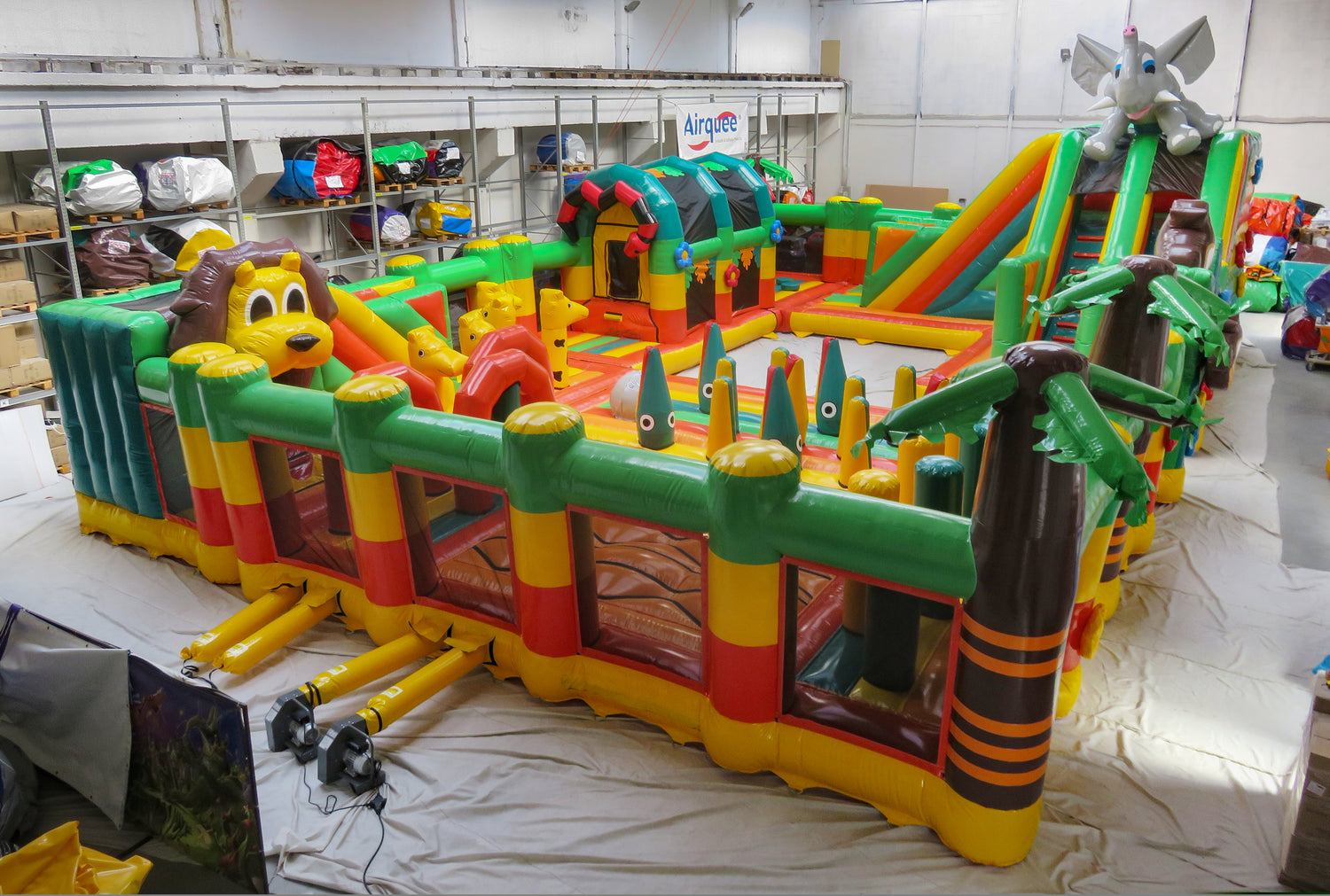 A large inflatable children's bounce house and slide set photographed in a warehouse. The colorful structure features green, yellow, red, and orange sections, cartoon animal faces, palm tree decorations, and a slide on the right side.