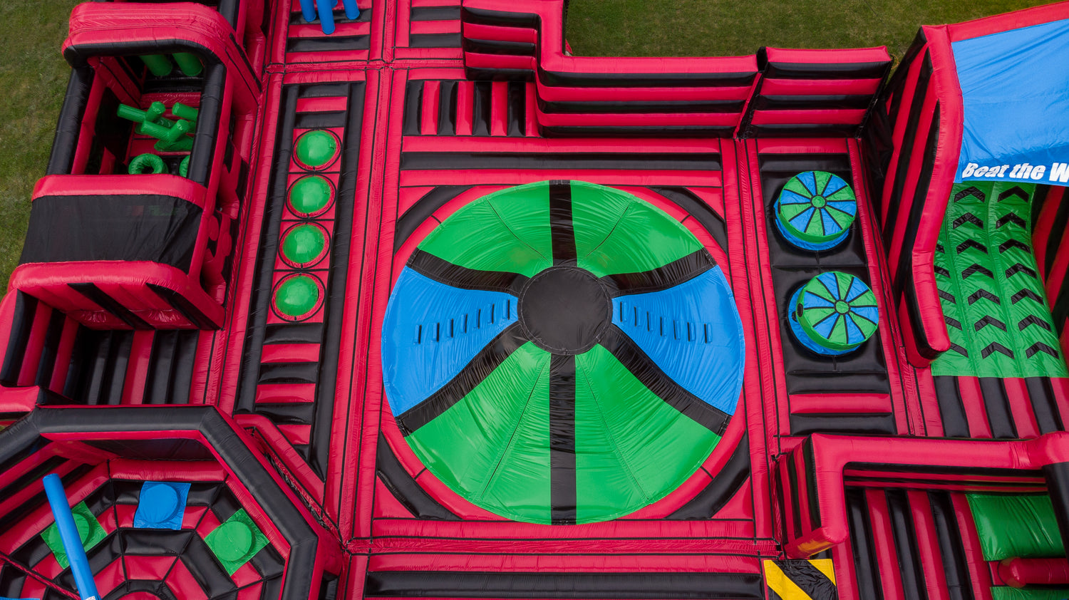 Aerial view of a large inflatable obstacle course or play structure with bright red, black, green, and blue sections, including a circular green and blue target area and multiple play zones.