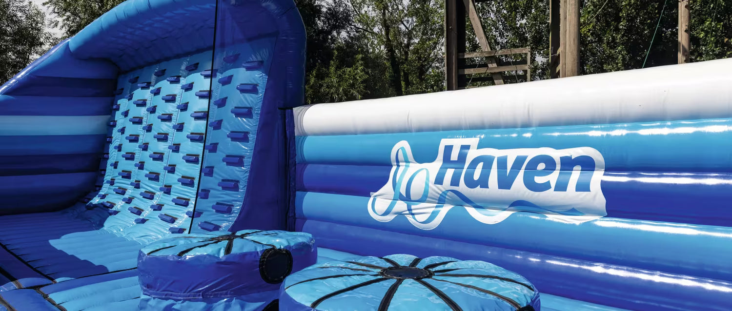 Blue and white inflatable pool play set with a slide, textured wall, and basketball-shaped pool float, branded with the Haven logo. Trees and outdoor structure are visible in the background.